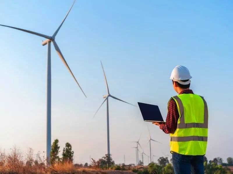 Man in high-vis carrying laptop looking at wind farm