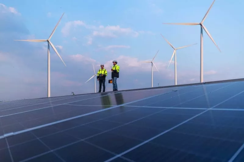 Two people standing on top of solar panels with wind turbines behind them